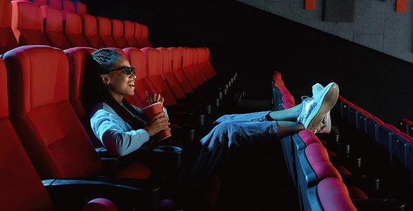 En ensam kvinna sitter i en biosalong med fötterna uppe på stolsraden framför och äter popcorn